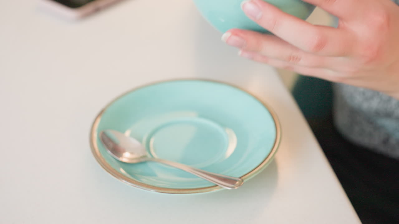 Close up of pretty fair lady seated gently sipping soft drink from ceramic cup on saucer with spoon relaxed cozy indoor setting visible smartphone on table warm natural light