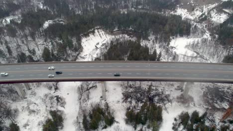 Toma-Aérea-De-Camiones-Que-Conducen-Sobre-Un-Puente-De-Carretera-Elevado-De-4-Carriles-En-Invierno-Lago-Congelado-Acantilados-De-Arena-Acantilados,-Terreno-Nevado-Paisaje-Forestal