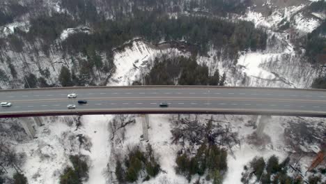 aerial tracking shot of cars driving over 4-lane elevated highway bridge in winter frozen lake sandy bluffs cliffs, snowy terrain forest landscape