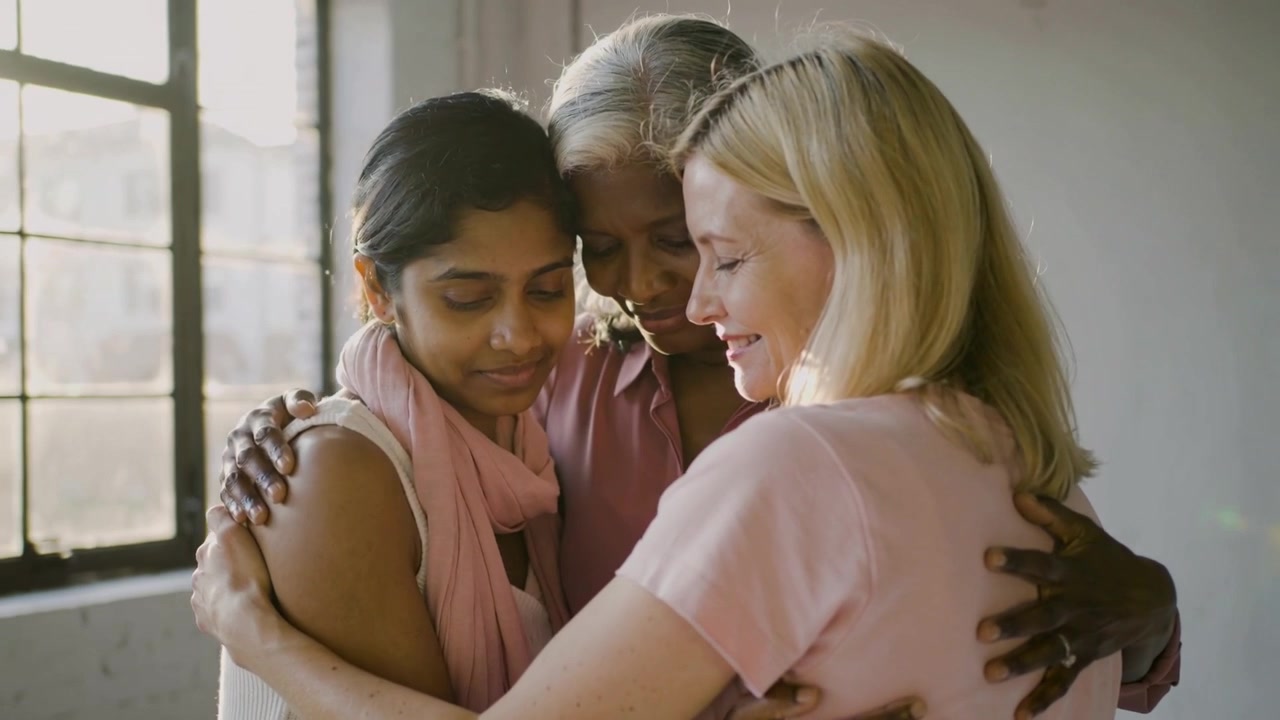 Ricongiungimento, tre donne che si abbracciano stretto davanti a una finestra luminosa, mostrando l'anello della nonna e una sciarpa rosa