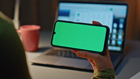 unknown lady holding greenscreen mobile phone at dark workplace laptop close up