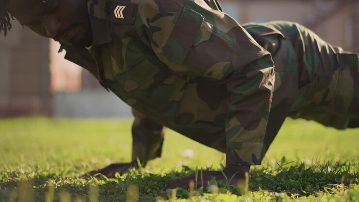 Esercitazione militare all'aperto, soldato disciplinato che si allena all'aperto in uniforme mimetica durante i piegamenti, personale militare concentrato impegnato in un allenamento di resistenza su un campo erboso con edificio sullo sfondo