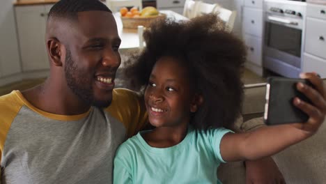 Hija-Afroamericana-Y-Su-Padre-Tomándose-Selfie-Juntos-Sentados-En-El-Sofá-Sonriendo