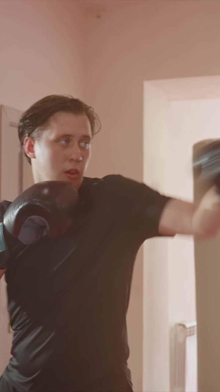 Giovane uomo in maglietta nera e guantoni da boxe si allena con i pugni. L'uomo mantiene la posizione di guardia e sferra jab e ganci. Ambiente interno luminoso con pareti chiare e telaio della porta.