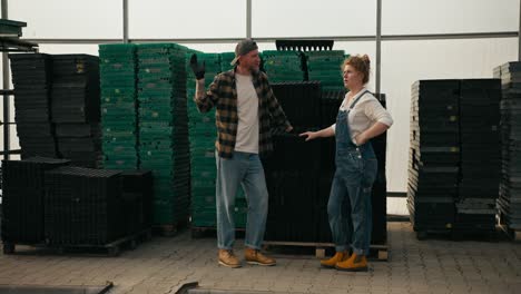 guy farmer with a beard in a plaid shirt communicates with his colleague a girl with red hair near seedling boxes in a greenhouse on a farm