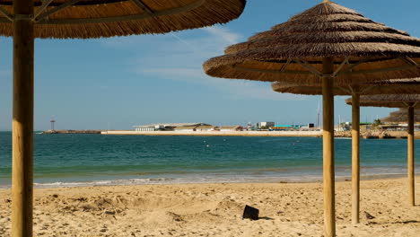 Empty-beach-with-waves-gently-lapping-at-the-shore,-view-under-thatch-umbrellas