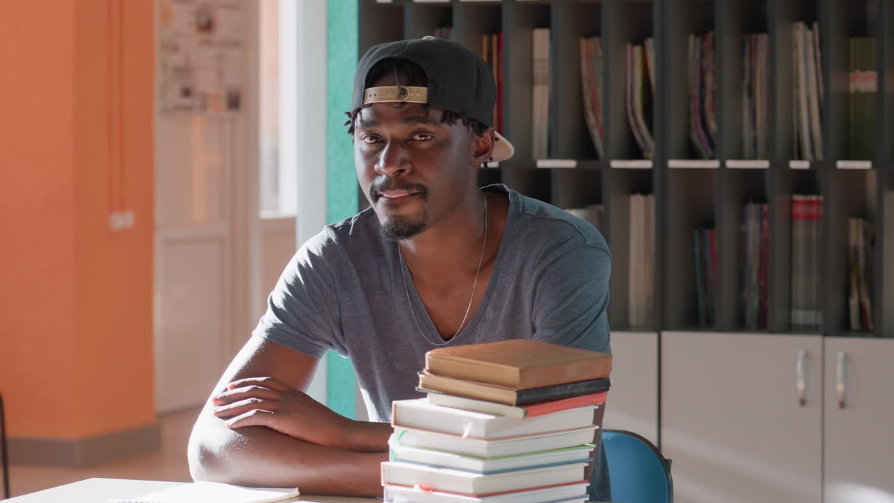 College student in cap sits at library table beside tall stack of books, arms resting and gazing ahead, ready for study session under warm light, shelves with magazines behind