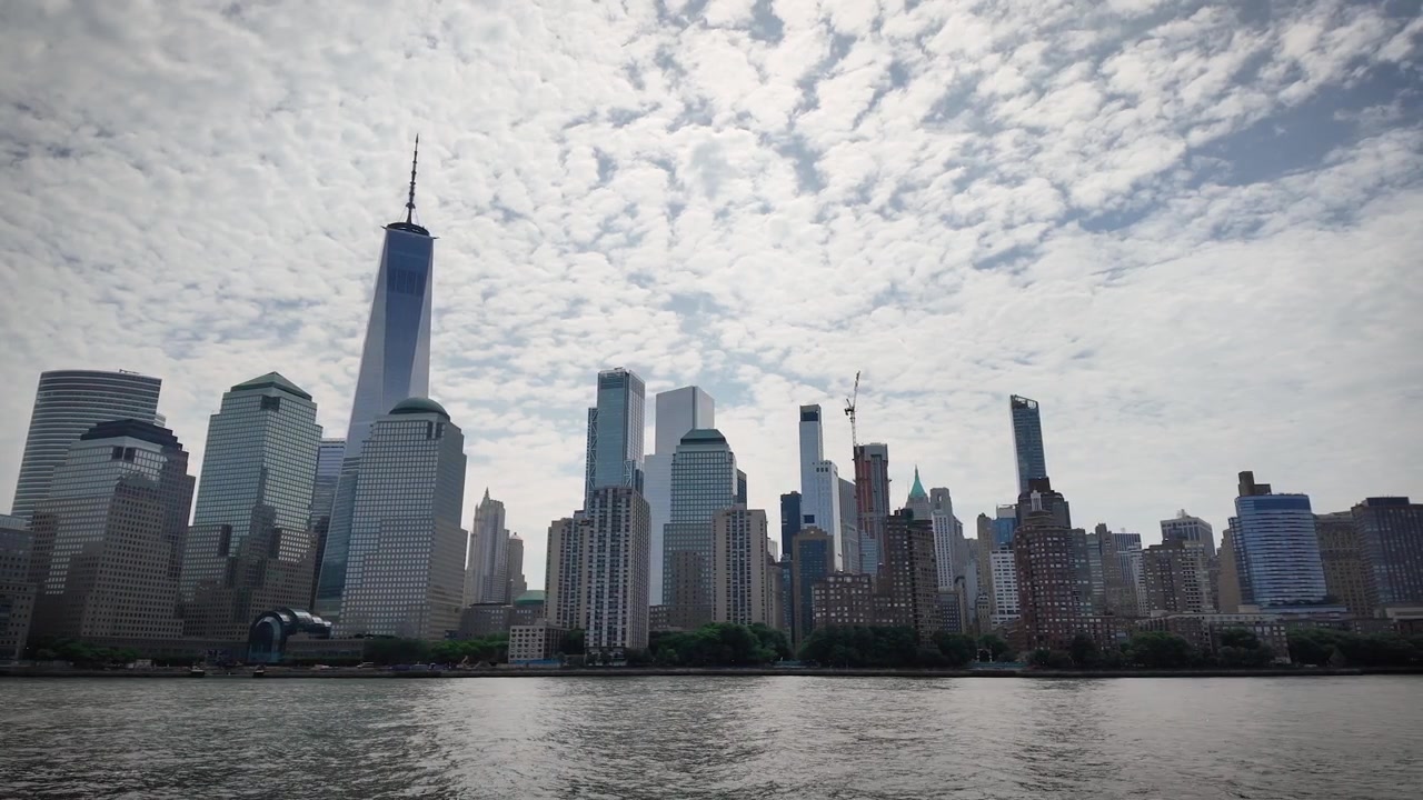 Ampia inquadratura cinematografica della Statua della Libertà su Liberty Island con acqua calma e nuvole drammatiche, vista dal porto di New York in una giornata limpida.