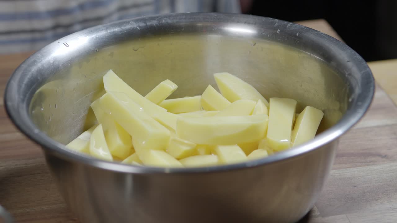 A chef pours olive oil over raw potato wedges in a metal bowl