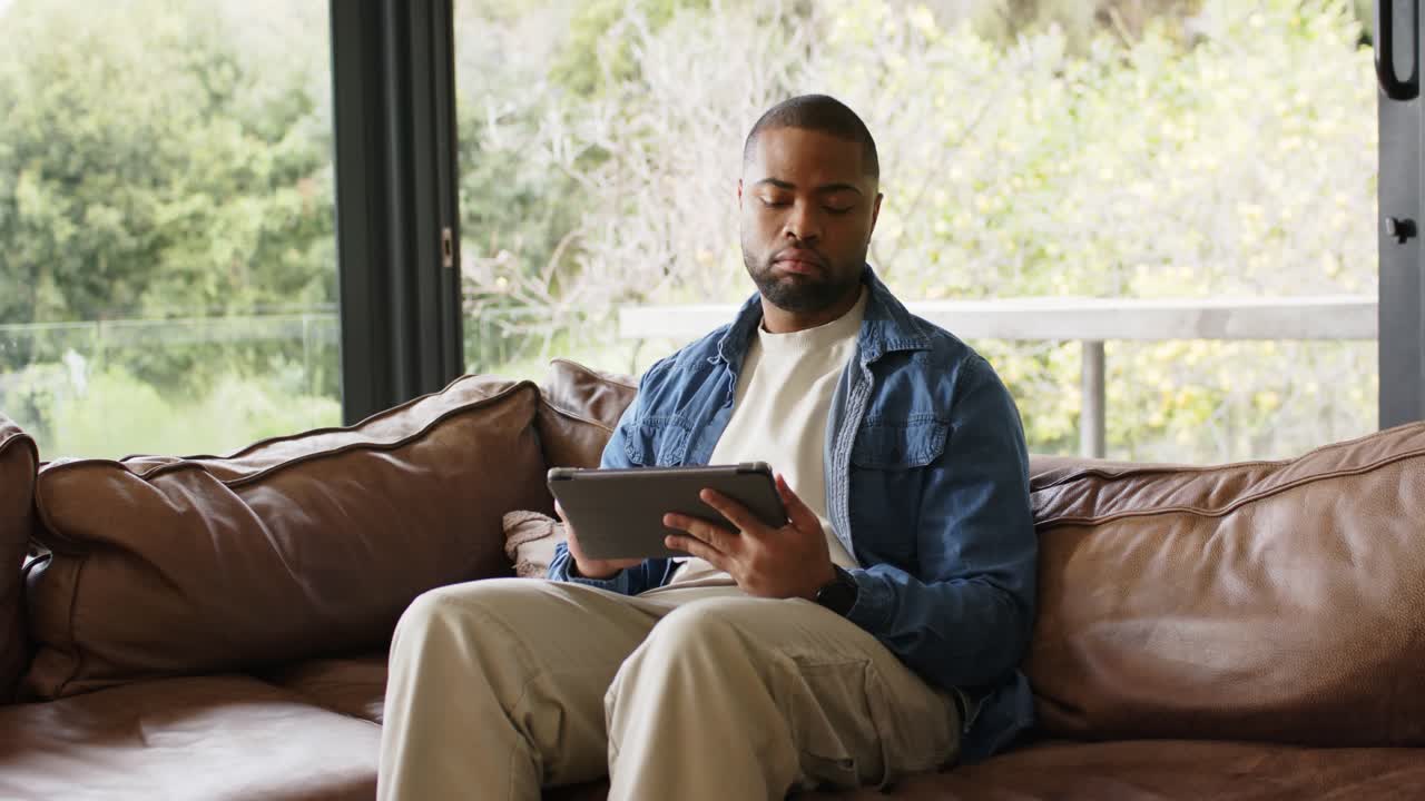 After propping tablet African American male tapping glancing outside reading on sofa by window