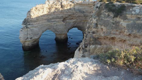 tilting up shot, scenic view of rock tunnel cliff in algarve, portugal, people standing on the top of the cliff on the bright sunny day