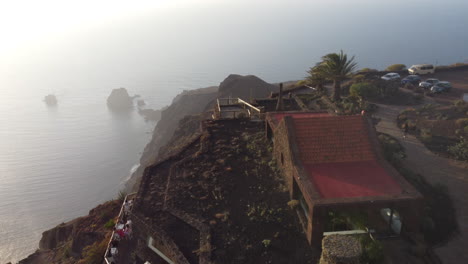 Un-Vuelo-Sobre-Un-Pequeño-Pueblo-Con-Edificios-Y-Turistas-Situado-En-Una-Montaña-Volcánica,-Debajo-De-La-Cual-Hay-Una-Costa-Con-Acantilados