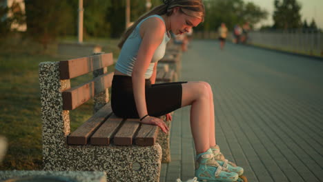 tired young lady sitting outdoors on a bench wearing roller skates, looking down with exhausted expression while dropping sneaker on ground, background features blurred greenery and people