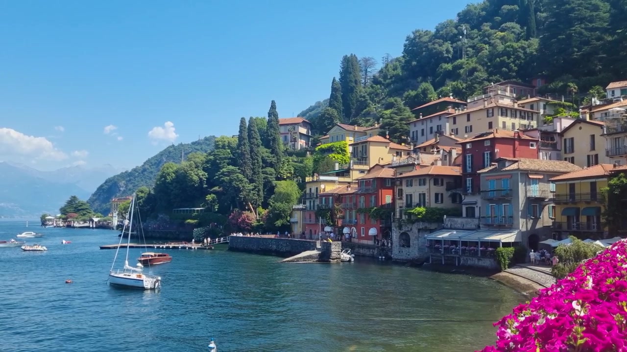 Una pittoresca scena estiva a Varenna sul Lago di Como, Italia, con piccole barche che riposano su calme acque alpine, vivaci fiori rosa e cieli azzurri che incorniciano questa iconica destinazione lacustre