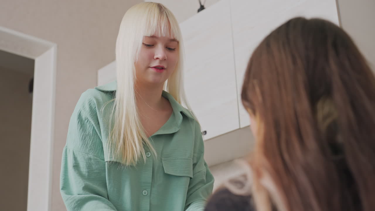 Blond tjej delar ett sött och kärleksfullt ögonblick med sin brunhåriga vän medan de leker i en köksmiljö, vilket uttrycker äkta samhörighet genom gester och uttryck under en rolig stund inomhus.