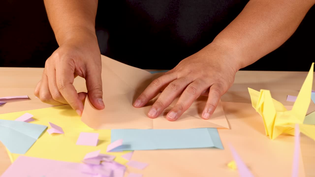 Person folds pastel origami paper into crane, overhead view, warm lighting, steady camera, creative mood