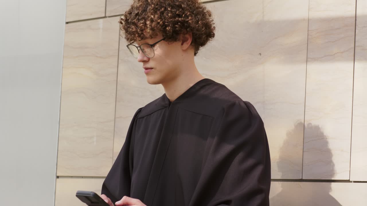 Formado do sexo masculino de beca perto de um muro de pedra olhando para cima e usando o smartphone ao lado do diploma, espaço para texto