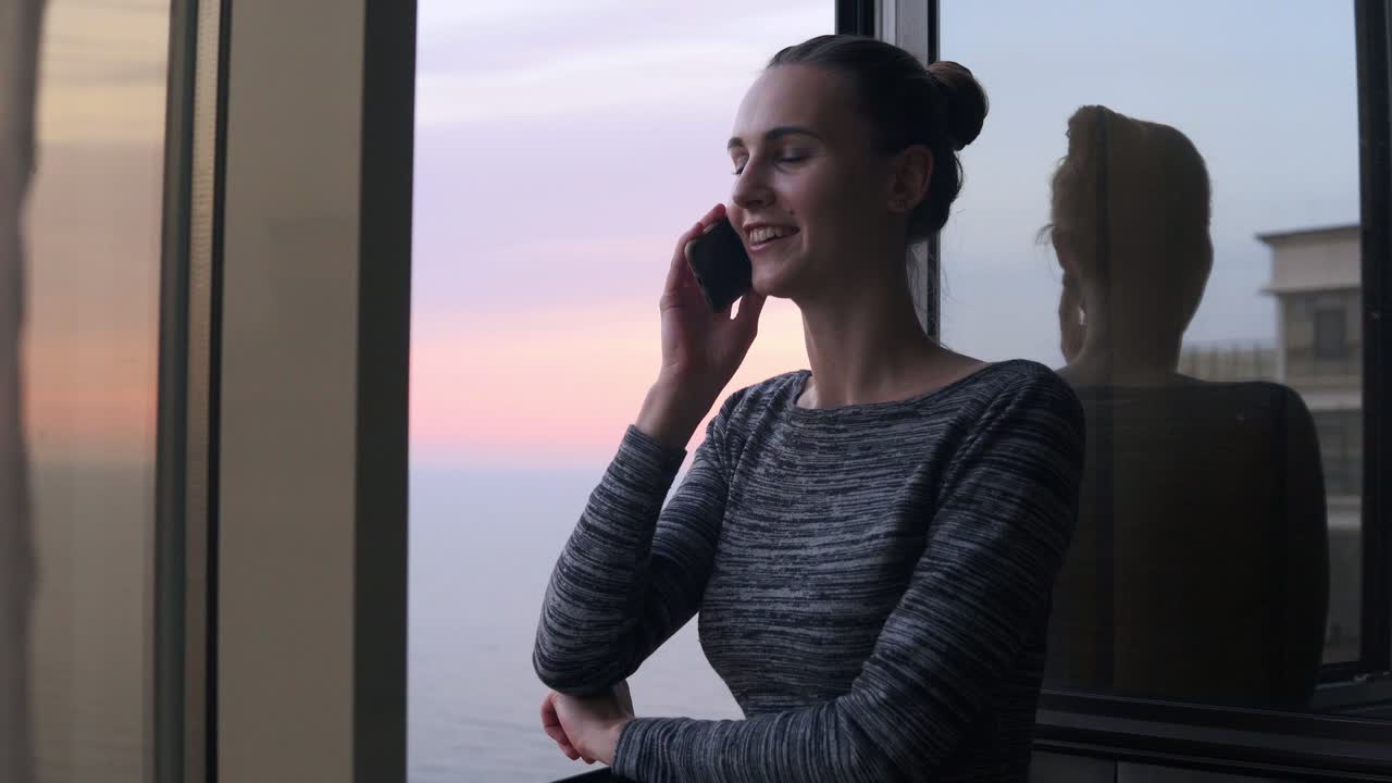 Young smiling woman dialing a number and starting to speak on the phone while standing by the open window with a smile during the sunset by the sea. Beautiful sky and sea on the background