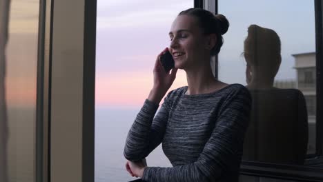 Young-smiling-woman-dialing-a-number-and-starting-to-speak-on-the-phone-while-standing-by-the-open-window-with-a-smile-during-the-sunset-by-the-sea.-Beautiful-sky-and-sea-on-the-background