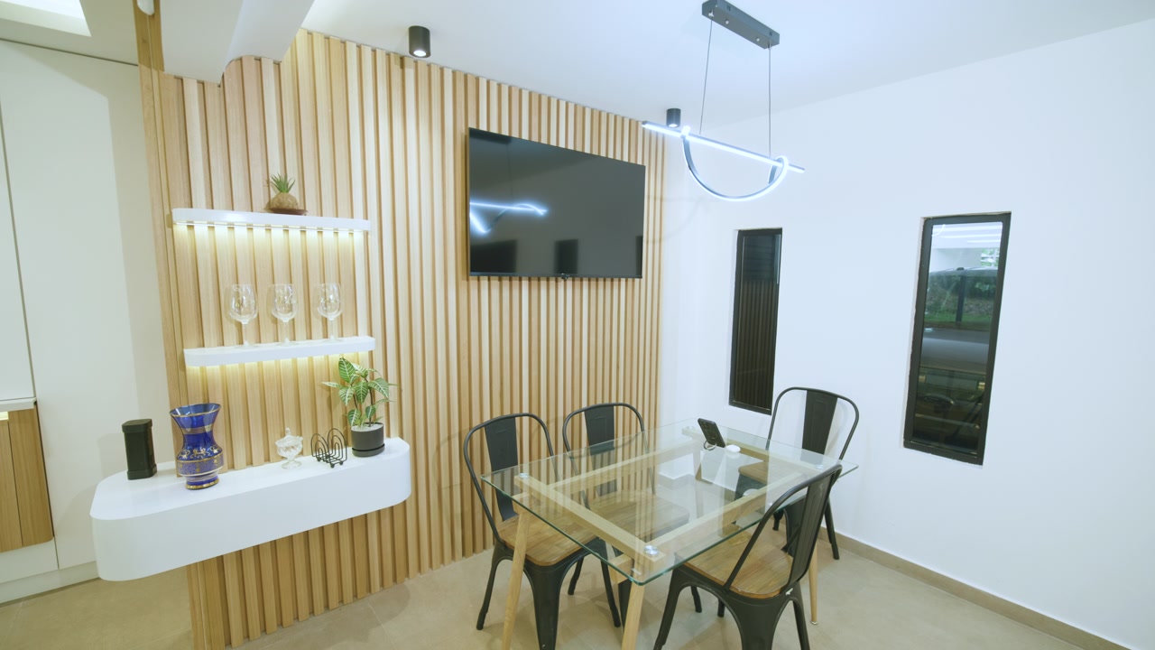 Full framing of the space: decorative wall with vertical wooden slats, illuminated shelves, side table, flat screen, dining table with chairs and designer pendant lamp
