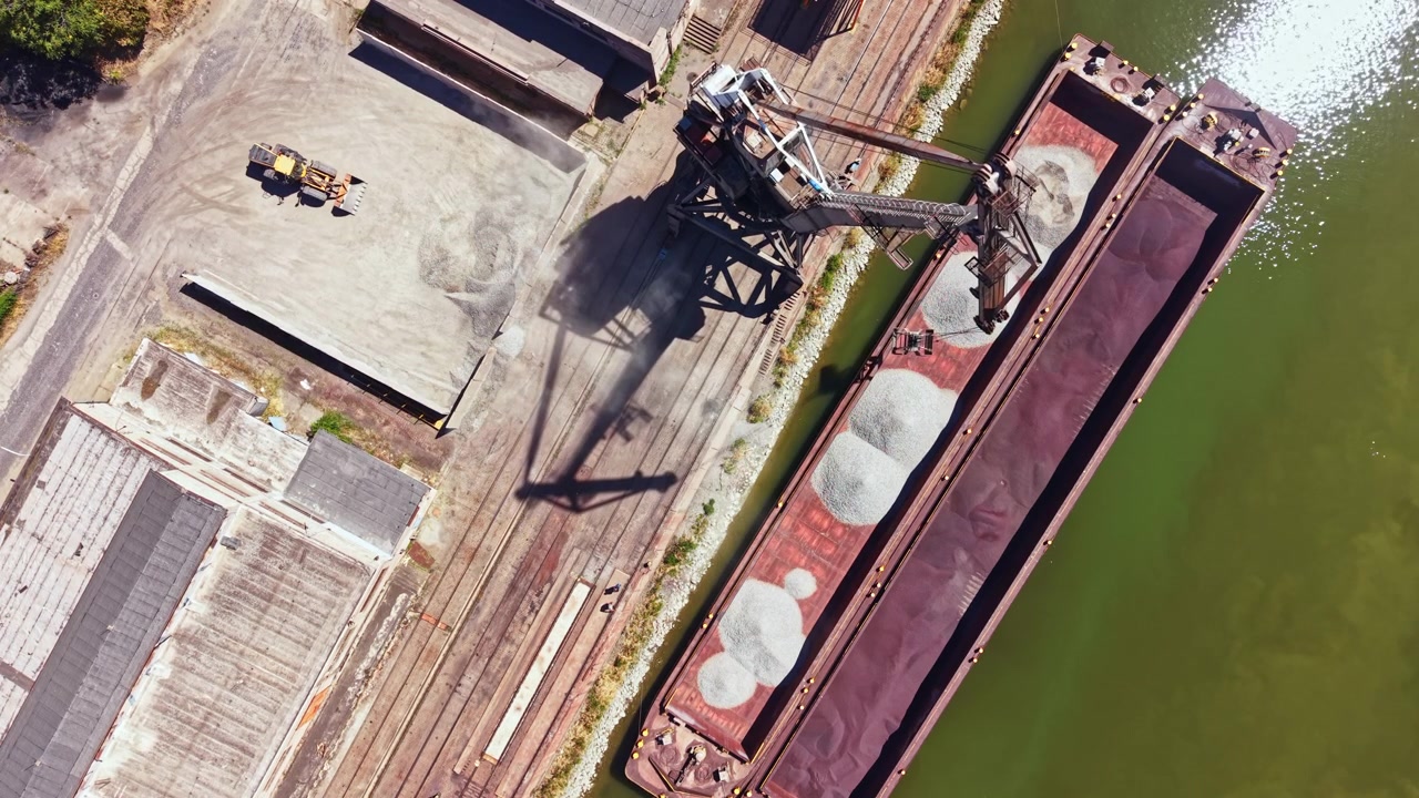 Carichi vengono trasferiti da un molo a una chiatta lungo un fiume in un'area industriale durante il giorno.