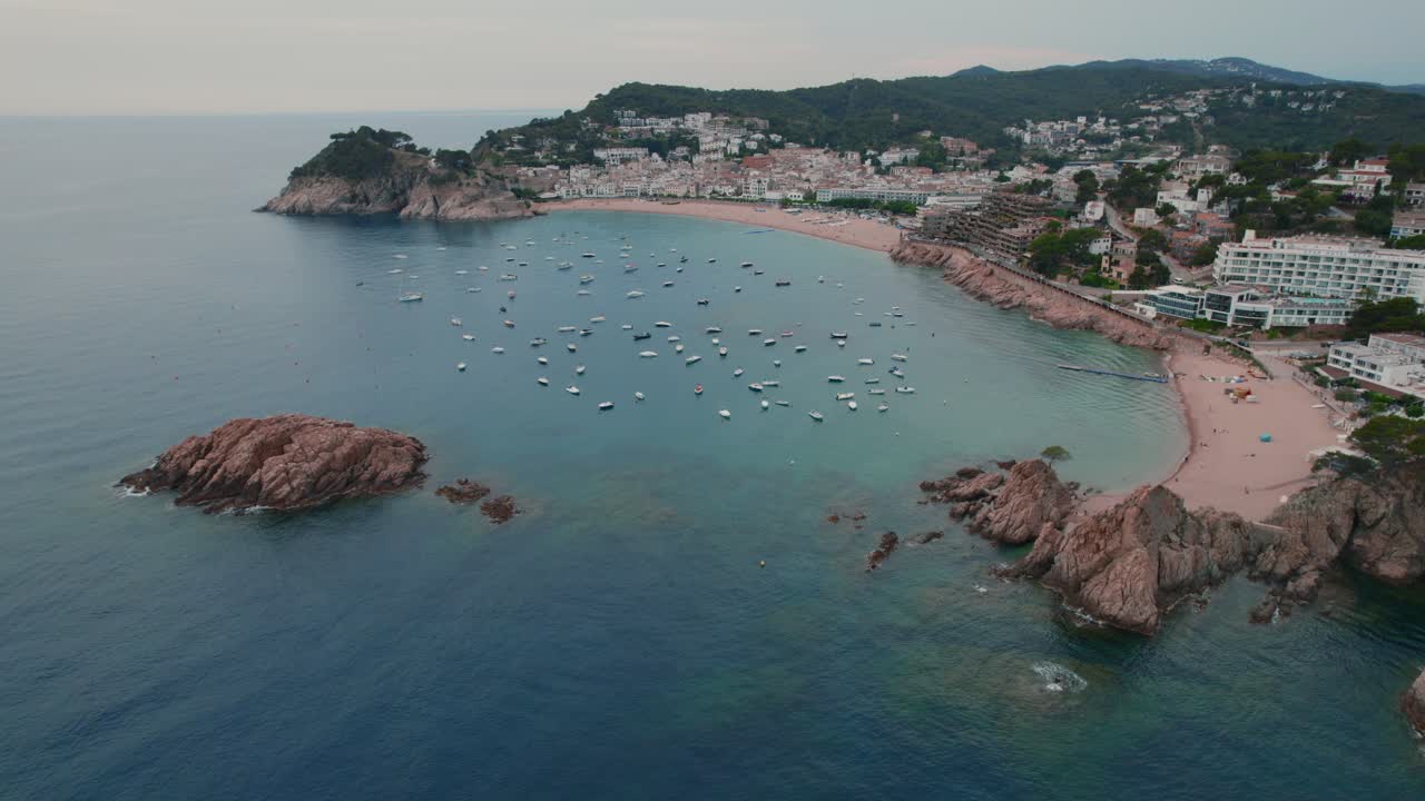 Sapuan kamera udara lebar Tossa del Mar di Costa Brava, Spanyol, dengan perahu-perahu di pelabuhan dan kota yang penuh kehidupan.