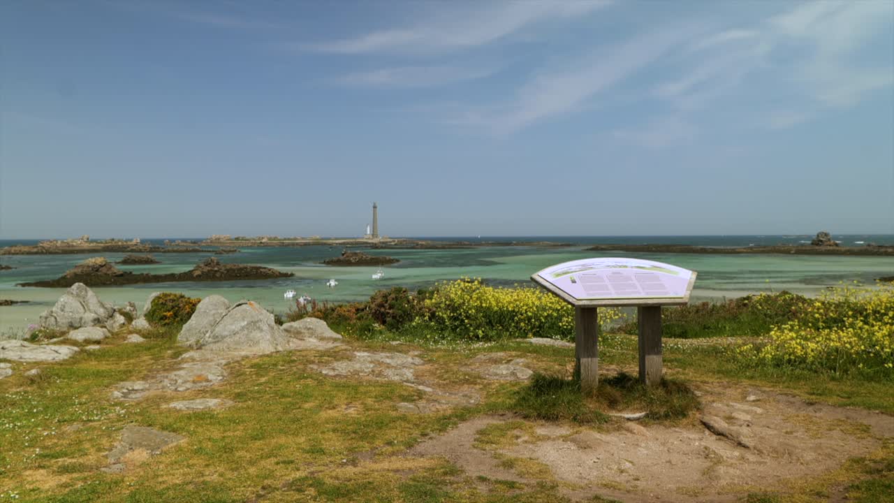 An information sign faces the Phare de l’Île Vierge lighthouse, set against a scenic landscape with yellow wildflowers, rocky shores, and boats in the clear turquoise water near Brittany, France