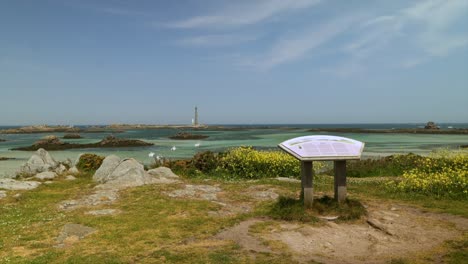 an information sign faces the phare de l’île vierge lighthouse, set against a scenic landscape with yellow wildflowers, rocky shores, and boats in the clear turquoise water near brittany, france