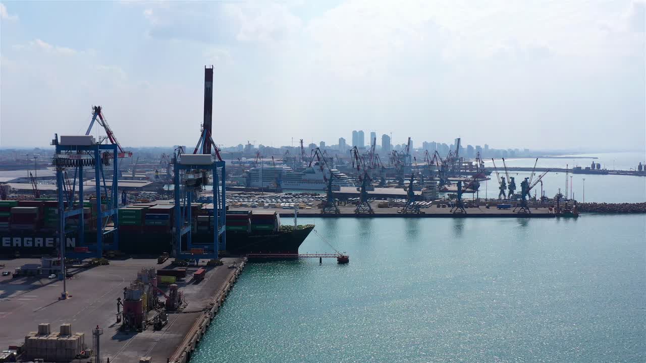 Aerial view of a large container ship docked at an bustling industrial port with cranes and a city skyline in the background