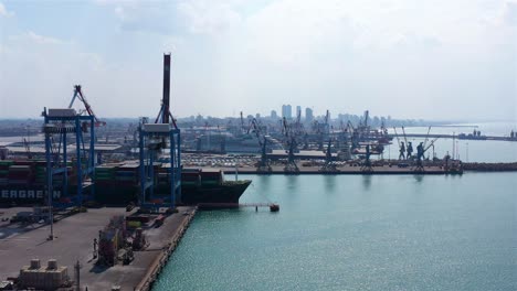aerial view of a large container ship docked at an bustling industrial port with cranes and a city skyline in the background