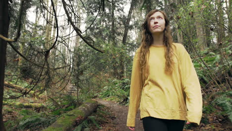 A-young-woman-wearing-a-bright-yellow-jacket-walks-through-a-green-mossy-forest,-tracking-shot-from-in-front