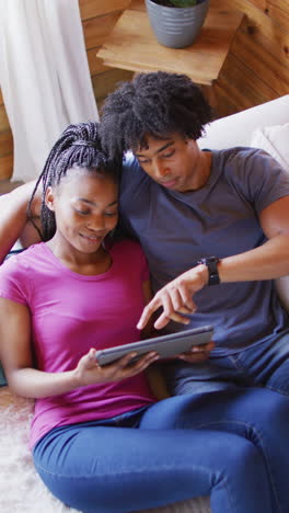 vertical video of african american couple sitting on sofa and using tablet in log cabin