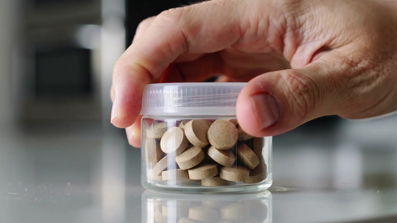 Hand entering top-right, right hand pressing and twisting lid on jar at counter, securing tablets
