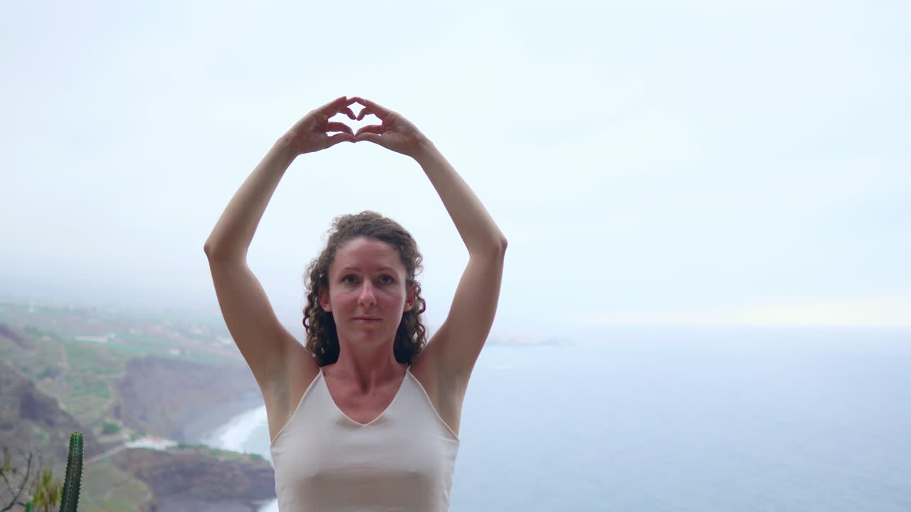 Amidst ocean and verdant mountains, a woman meditates atop a peak, her hands forming the Maha Sakal gesture