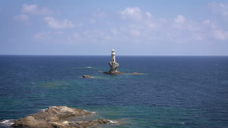 solitary lighthouse stands on a small rocky island in the middle of the blue sea, with distant hills under a cloudy sky