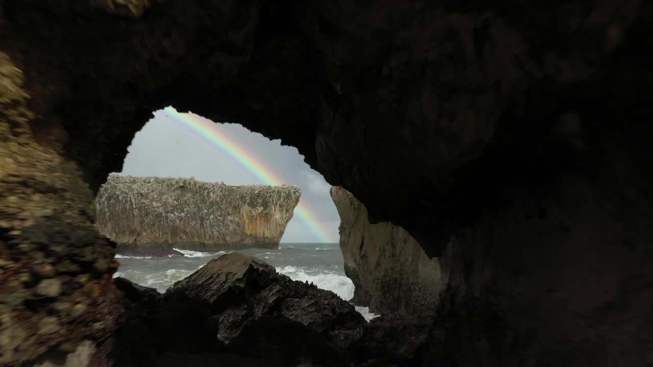 vista aérea de drones a través de un arco, revelando un arco iris en la costa de la playa de las cuevas, en el norte de españa