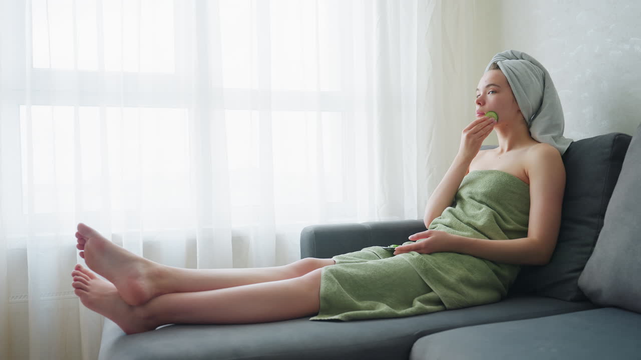 Woman relaxing indoors, sitting on couch wrapped in light green towel, gently massaging cheeks and jaw with cucumber slices for skincare in bright, serene space filled with soft natural daylight