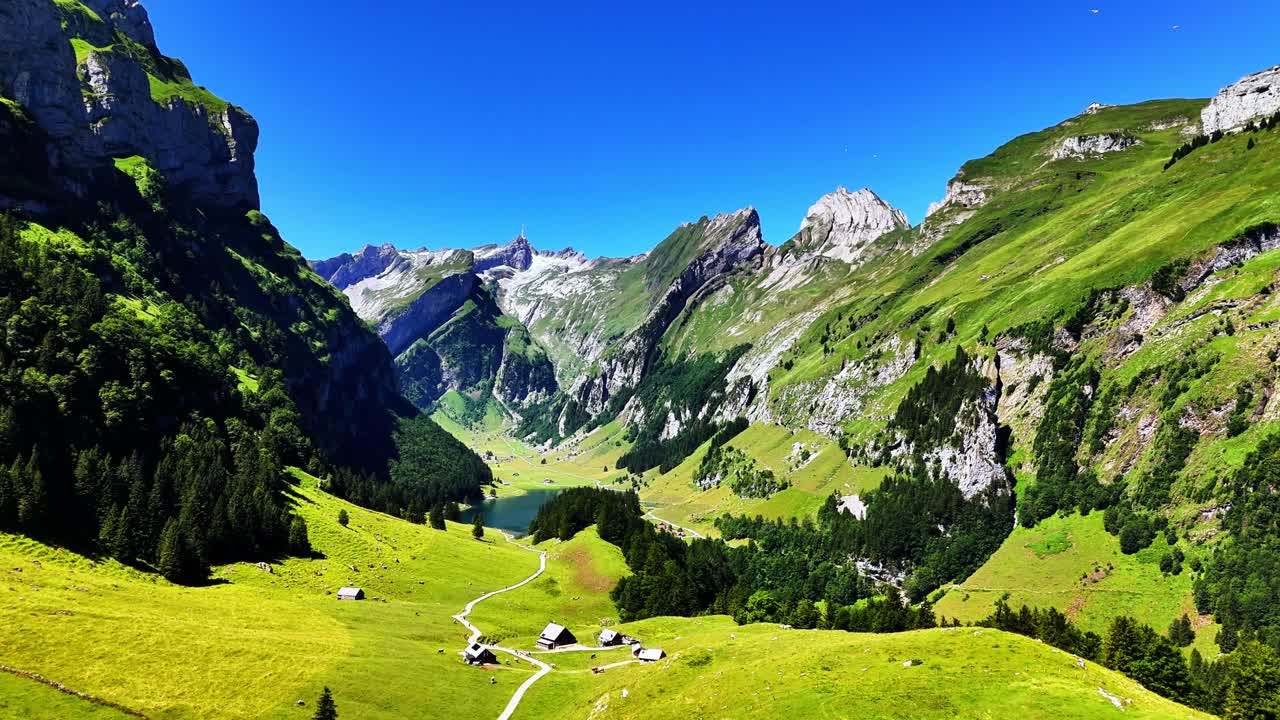 Naturskjønn utsikt over Seealpsee i Sveits med grønne enger, alpine hytter, skogslier, bratte fjellklipper, svingete stier og en klar innsjø under en klar, blå sommerhimmel