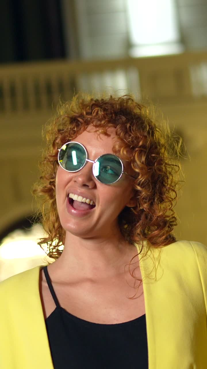 Donna con capelli rossi ricci che indossa occhiali da sole e un blazer giallo