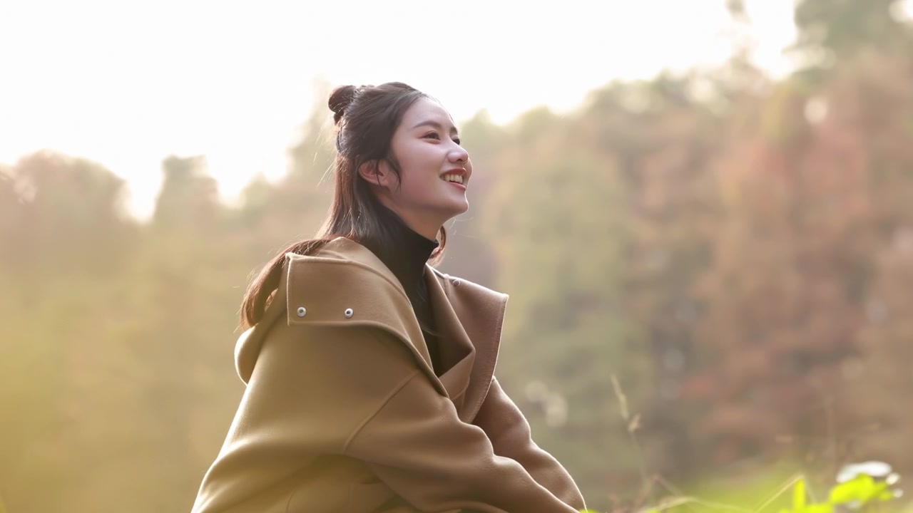 冬の装いをした陽気な若い観光客の女性が、自然豊かな秋の公園で落ち葉と楽しんでいる。晴れた日の朝の背景に、金色の紅葉の中で屋外の秋の季節を楽しむ笑顔の女性。