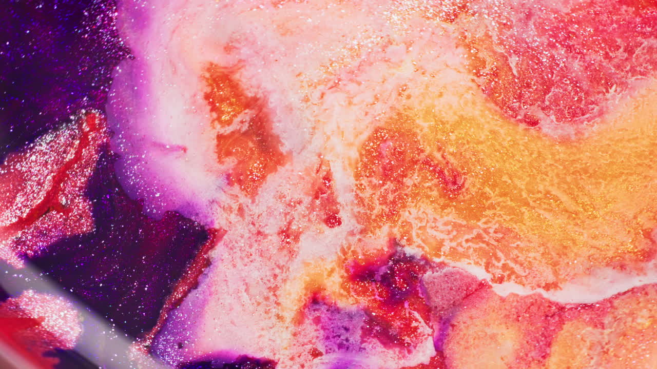 Macro shot of purple, orange, and pink glitter inks reacting to an air injection, forming a central void and shifting fluid particles