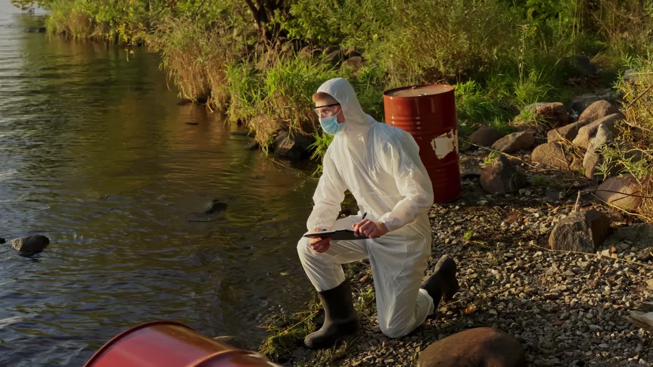 Un operatore ambientale in tuta protettiva esamina un barile rosso sulla riva del lago mentre valuta un possibile inquinamento