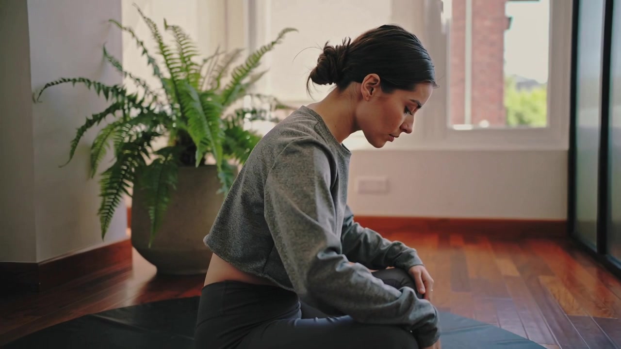 Donna che espira e si piega in avanti sul tappetino a casa, indossando un crop top e leggings, vicino a una felce per la centratura.