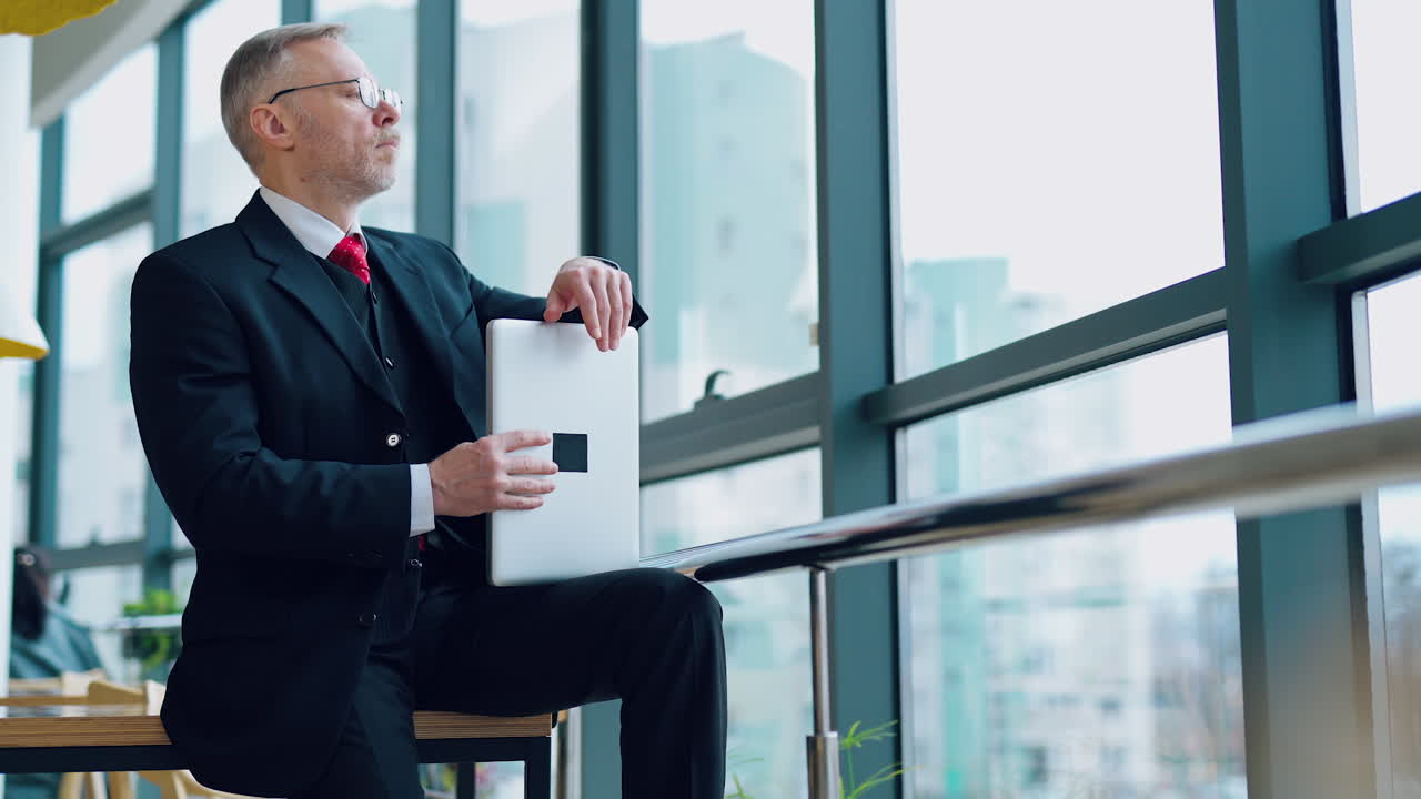 Serious entrepreneur in suit in office. Mature businessman sitting on the table with a laptop and looking through the window thoughtfully.
