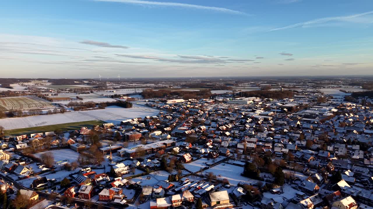 Vista aérea de uma cidade alemã coberta de neve durante o inverno, com luz solar baixa iluminando telhados, bairros residenciais e campos circundantes sob um céu azul claro. Foto ampla