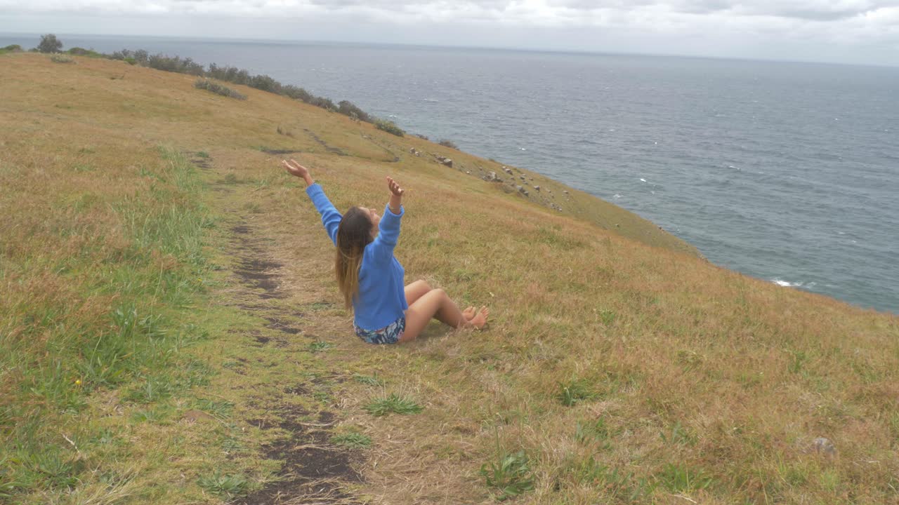 mujer joven disfrutando de la vista al mar levantando las manos mientras se sienta en una colina cubierta de hierba en el mirador de crescent head en nsw, australia