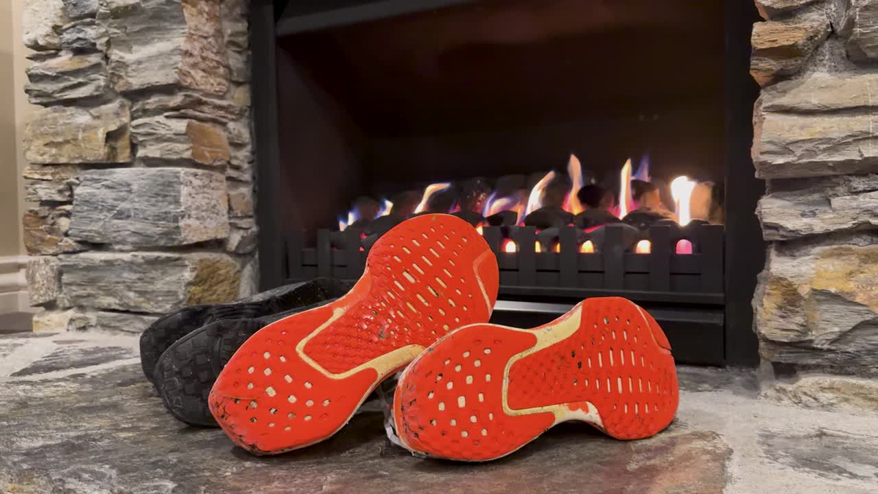 A pair of hiking shoes with orange soles dries in front of a lit stone fireplace. Warm, steady lighting and static camera create a cozy mood