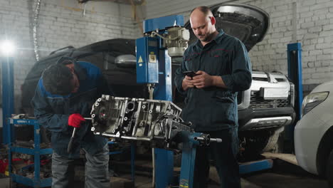 automobile engineers in a workshop, one intently working on an engine while the other is engaged with his phone, illuminated by a bright light in the background