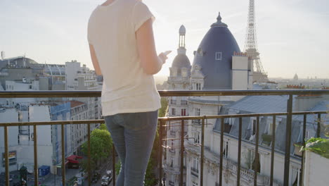 Glückliche-Frau,-Die-Ihr-Smartphone-Nutzt-Und-Auf-Dem-Balkon-In-Paris,-Frankreich,-SMS-Schreibt-Und-Den-Blick-Auf-Den-Eiffelturm-Genießt-Und-Ihr-Urlaubserlebnis-Beim-Surfen-In-Den-Sozialen-Medien-Teilt.-Wunderschöner-Sonnenuntergang