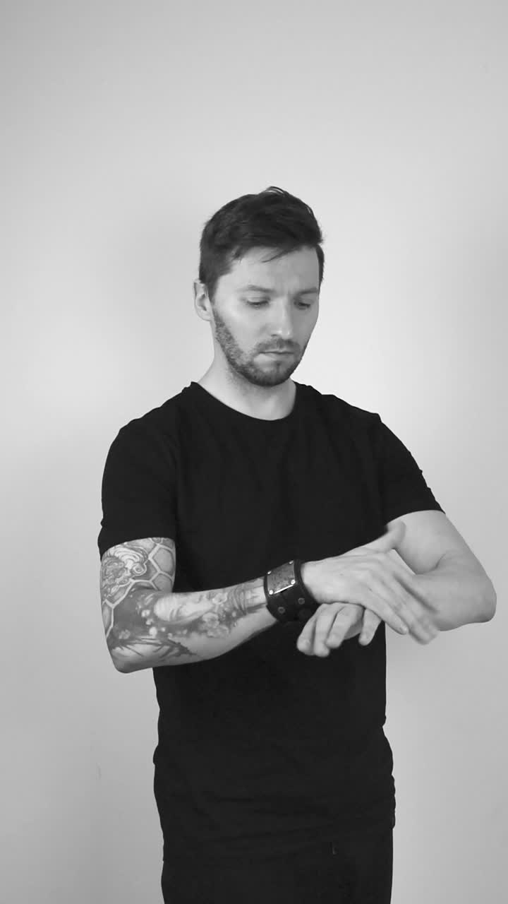 Grayscale footage of a white tattooed man with short beard and hair flipping a Bitcoin cryptocurrency coin in the air making it spin and hover, then catches it and puts it on his arm to check result.